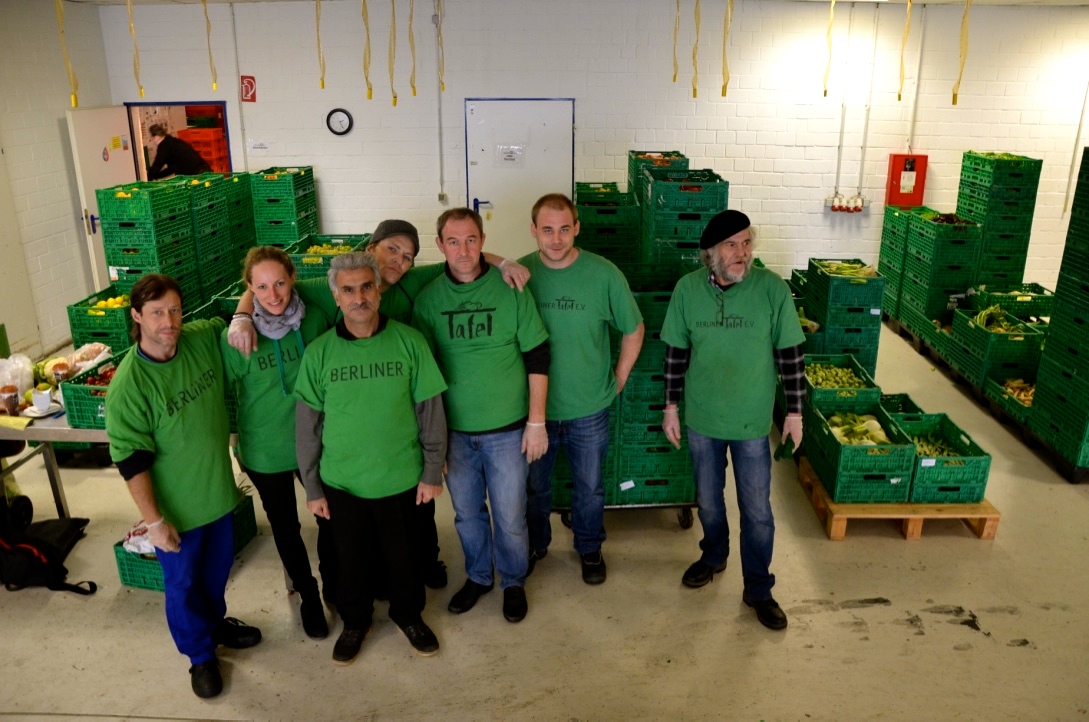 Sieben Personen in grünen T-Shirts bei der Berliner Tafel mit grünen Kisten voller Lebensmitteln hinter ihnen