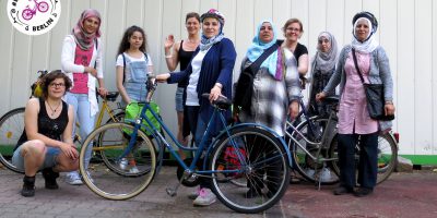 Mehrere Frauen stehen beisammen, einige mit Fahrrädern und Helmen, oben links im Bild das Logo der Organisation Bikeygees