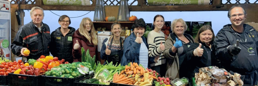 8 Personen unterschiedlichen Alters stehen hinter einen Tisch voller frischem Gemüse, das an Bedürftige verteilt werden soll