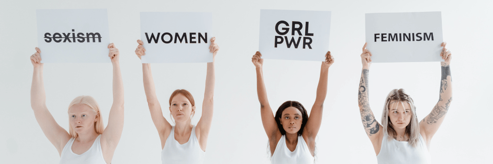 4 Frauen halten Schilder mit Begriffen hoch: Sexism (durchgestrichen), Women, GRL PWR, Feminism
