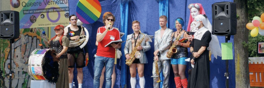 Musiker*innen stehen auf einer Bühne in bunten ausgefallenen Kleidungsstücken beim queeren Fest von AHA