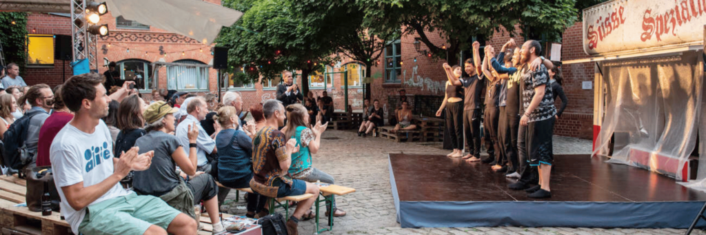 Schauspieler*innen auf einer Bühne verbeugen sich vor ihrem Publikum auf einer kleinen Bühne in einem großen Hof zwischen gemauerten Gebäuden.