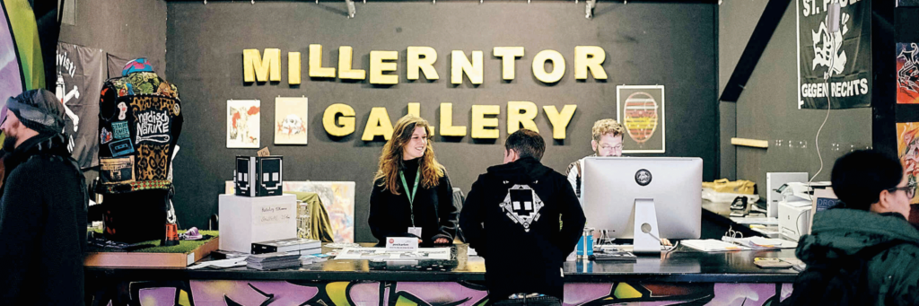 Einlass der Millerntor Gallery mit 2 Personen hinter dem bunten mit Graffiti gestalteten Tresen im Gespräch mit einem Besucher