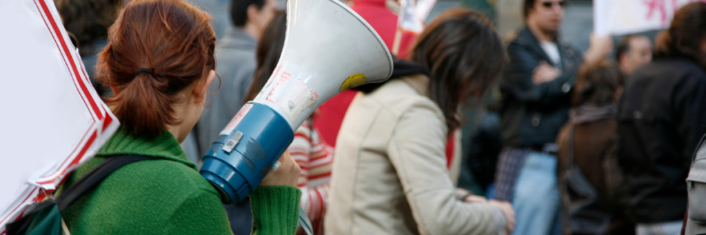 A woman can be seen from behind. She is holding a sign and a megaphone.