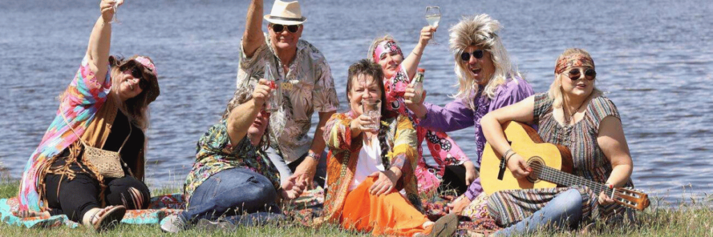 Eine Gruppe Menschen sitzen im Gras am See und feiern mit bunten Klamotten und Gitarre. 