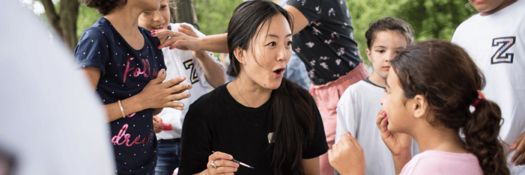 An adult woman, surrounded by children, whom she appears to be putting make-up on.  