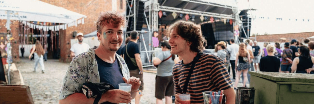 Zwei Menschen unterhalten sich und lachen dabei. Im Hintergrund sind andere Menschen und und ein Festivalgelände mit Bühne zu sehen.  