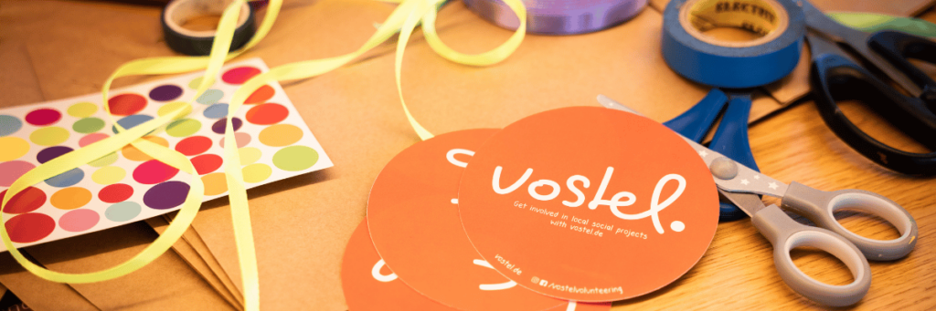 On a table are craft items such as scissors, colorful strings and stickers.
To the right of the center is a round, orange sticker with the vostel logo.   