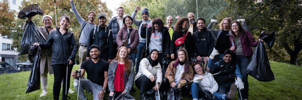 A group photo of many people who have just helped with a clean-up campaign. Everyone smiles happily at the camera and holds up the garbage grabbers and the filled garbage bags. 