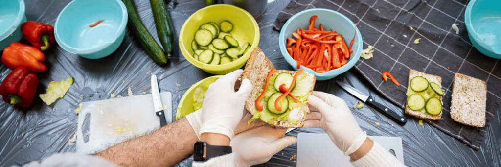 A bread roll is being filled.
On the table in the background are bowls of cucumber and peppers.   