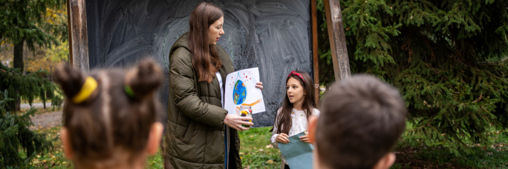 Frau erklärt Kindern in einem Wald, Dinge über den Klimaschutz, anhand eines Plakates.